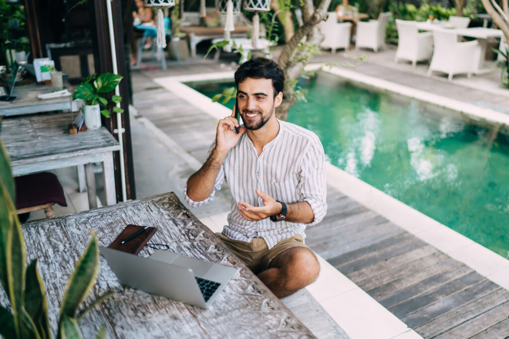 Man on phone with laptop by a pool. Looking for the best travel insurance for digital nomads